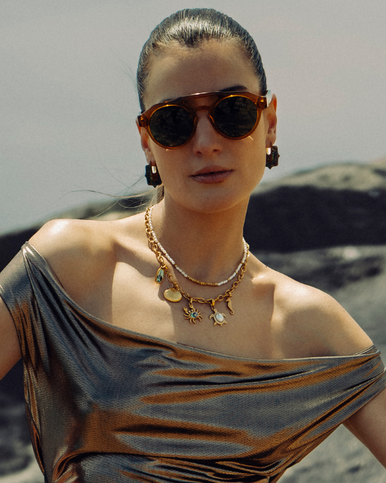 Woman in sunglasses wearing layered gold and pearl necklaces by the sea